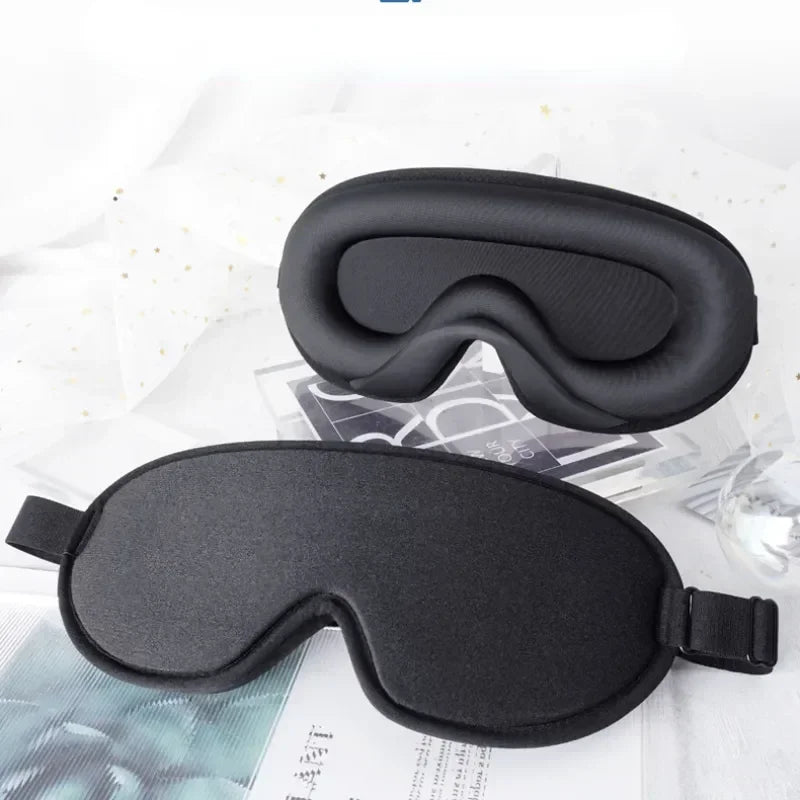 Black contoured 3D sleep mask with its adjustable flat strap, on white bedding