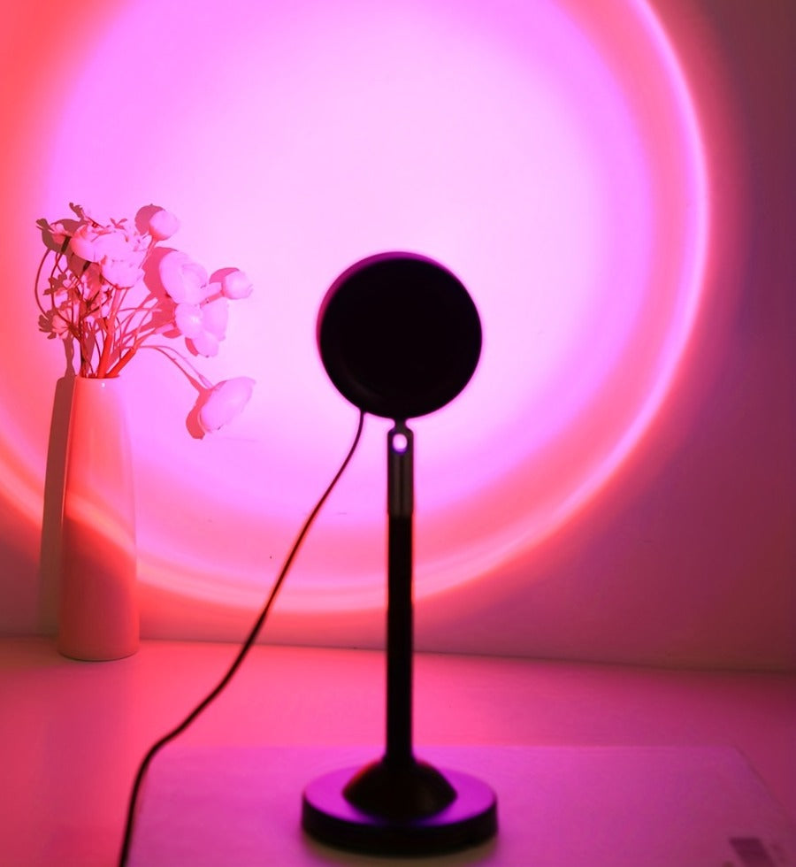 Sunset projection lamp on its stand casting a pink glow on the wall beside a vase of flowers